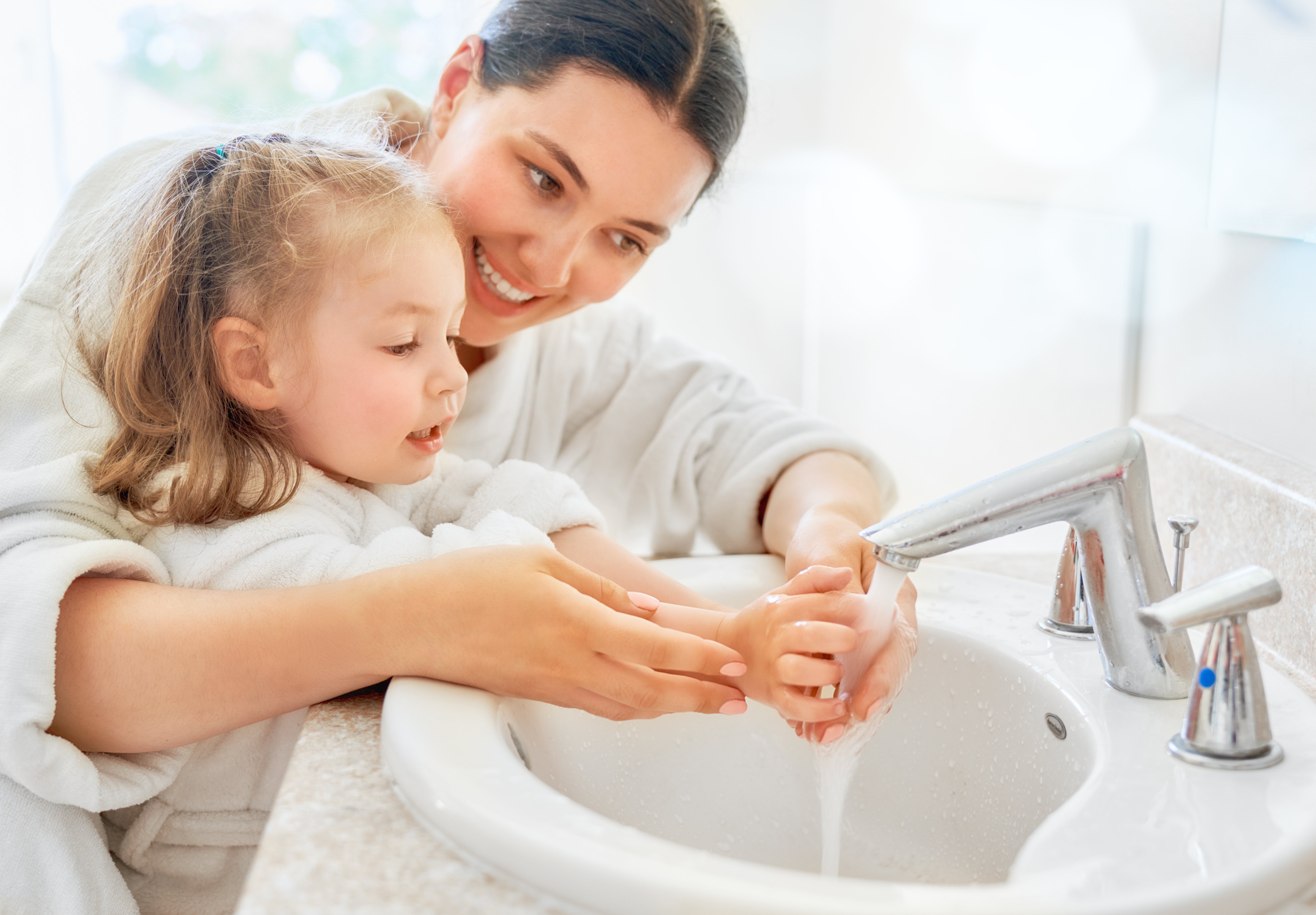 A parent washing hands at a bathroom sink while a preschool-aged child watches nearby, realistic photo