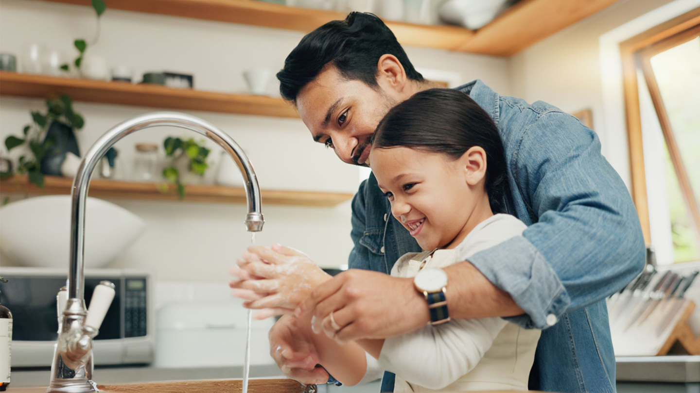 A parent washing hands with soap at a kitchen sink with a toddler watching from a safe distance, realistic photo