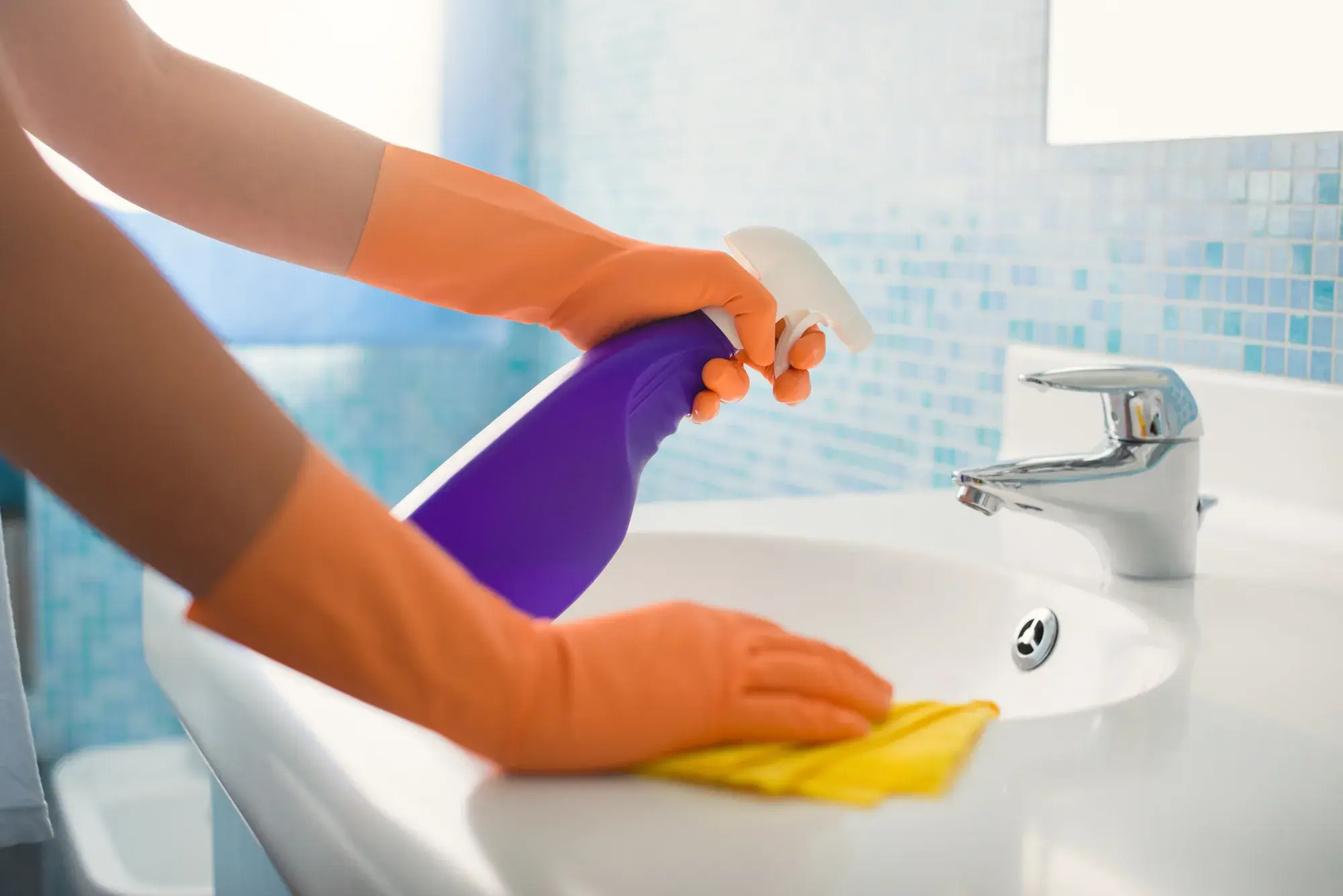 A parent wearing disposable gloves wiping down a bathroom sink and faucet with disinfectant, realistic indoor photo