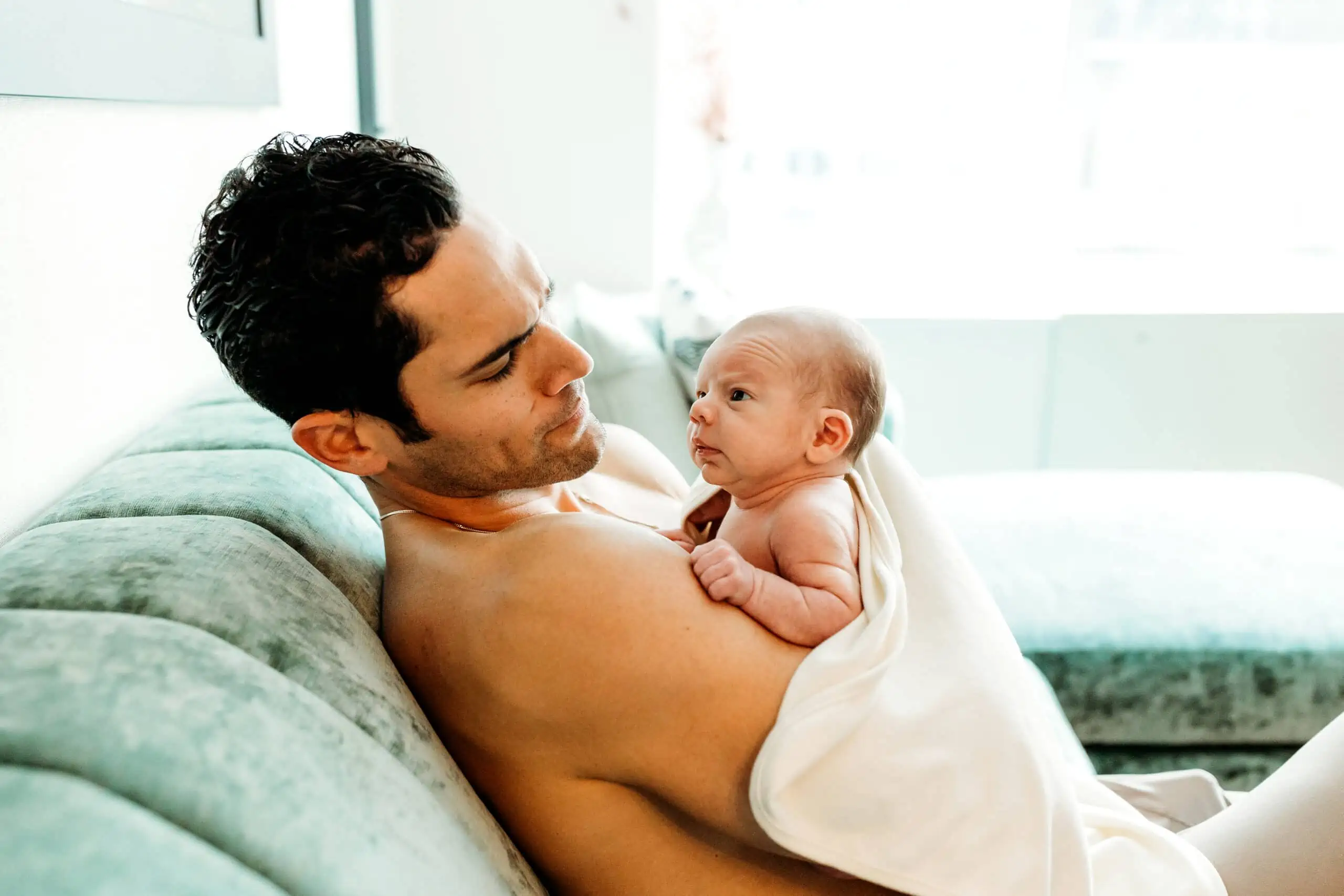 A partner sitting at a kitchen table with a phone and notepad while a postpartum parent rests nearby on the couch with a blanket, showing practical support and care