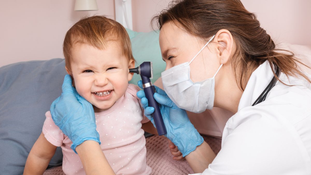 A pediatric ENT doctor examining a young child in a clinic room while a parent sits nearby, real medical visit photograph