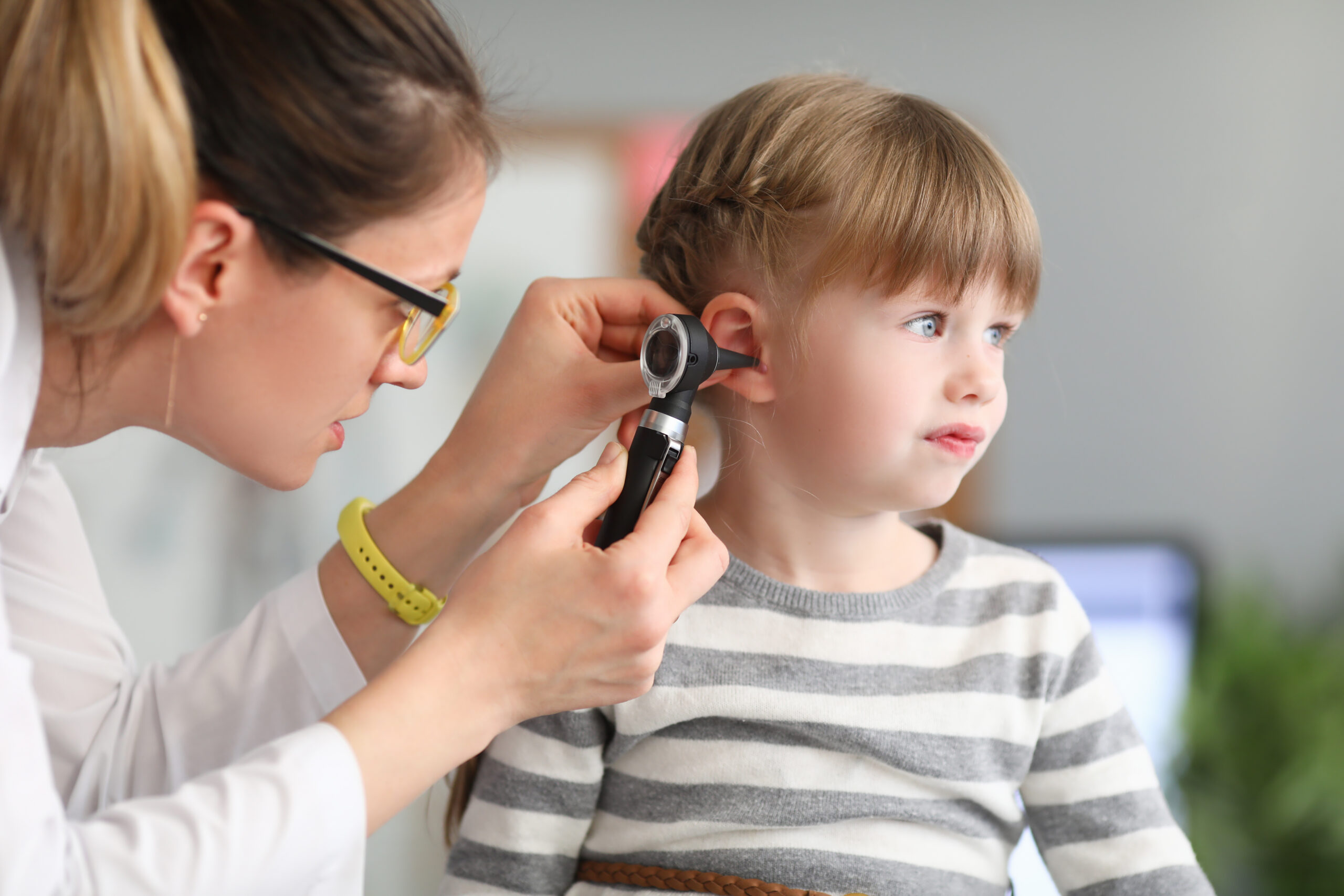 A pediatric clinician gently examining a preschool-aged child’s ear with an otoscope in a well-lit clinic room while a parent sits nearby, realistic medical photo