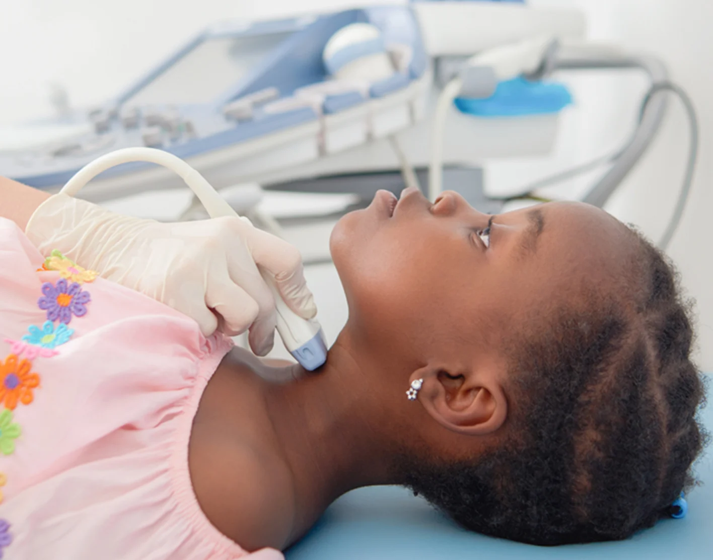 A pediatric clinician performing an ultrasound scan on a child's neck in a medical exam room with soft clinical lighting