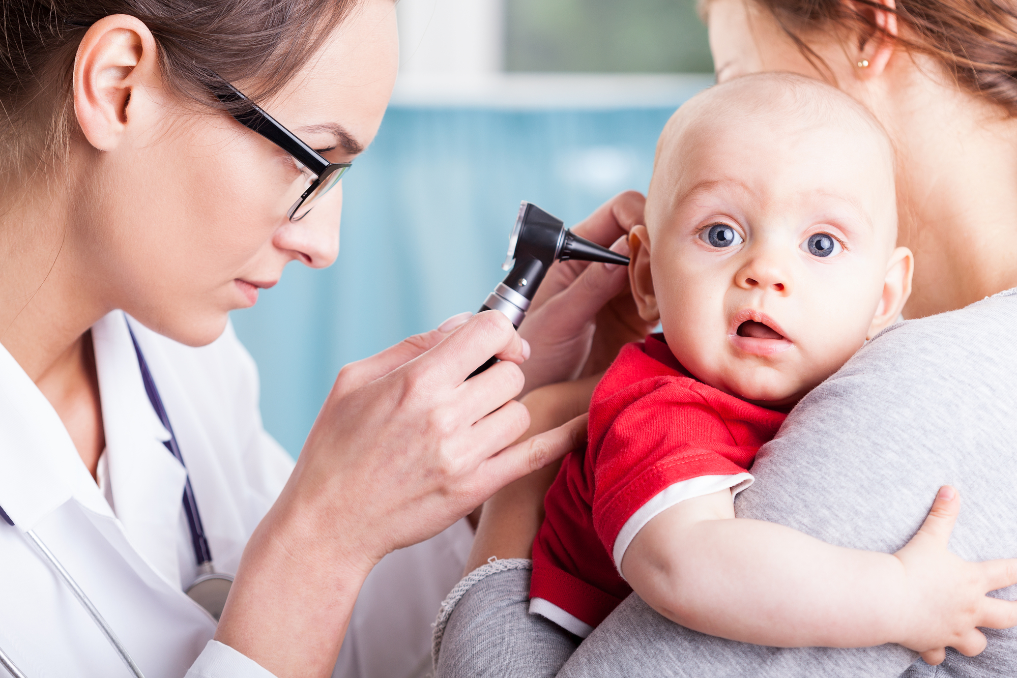 A pediatric clinician using an otoscope to look into a child’s ear while the child sits on a parent’s lap in a bright clinic room, realistic medical photo