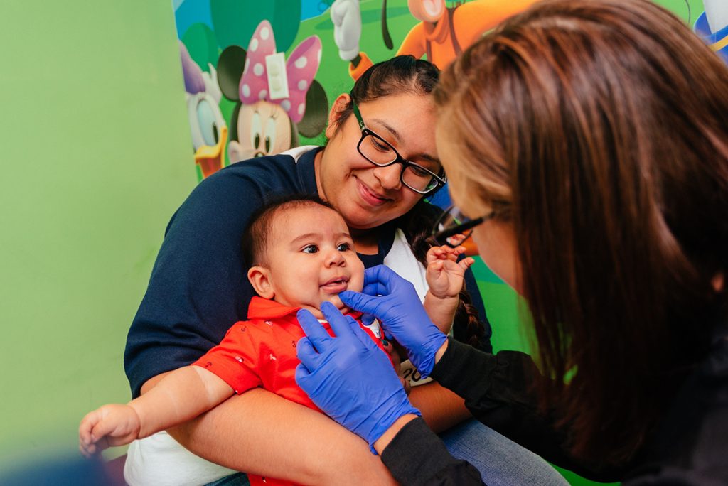 A pediatric dentist gently examining a baby’s mouth while the baby sits on a parent’s lap in a bright dental office