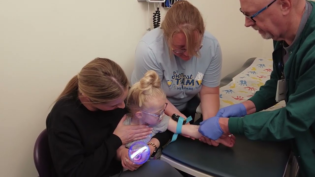 A pediatric nurse gently preparing supplies for a toddler blood draw in a clinic room while a parent comforts the child, candid healthcare photo style