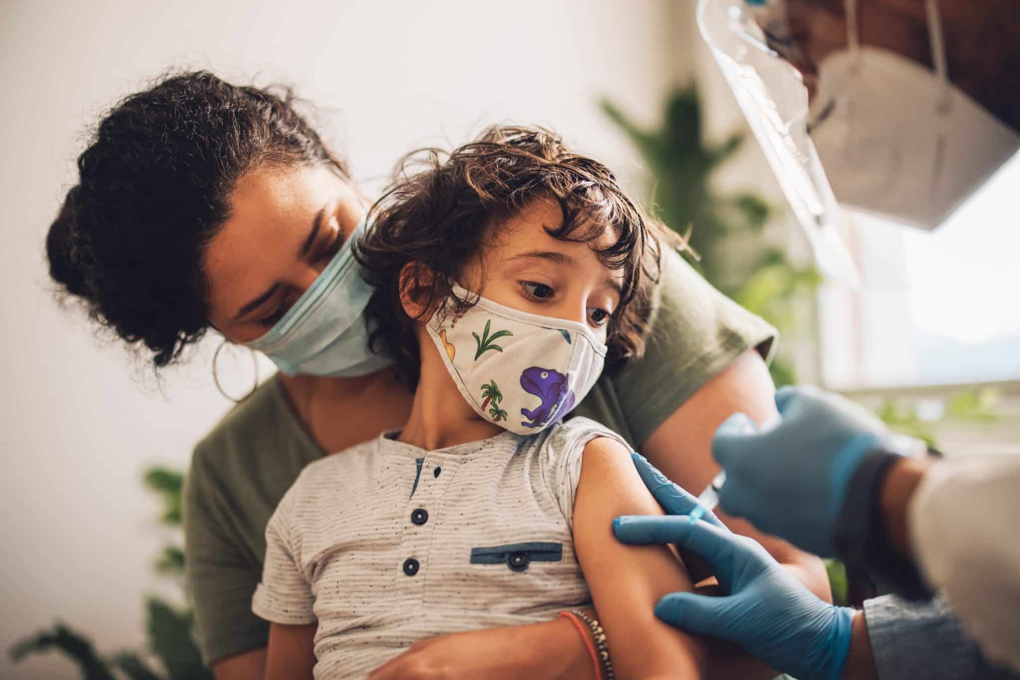 A pediatric nurse preparing a flu vaccine syringe while a parent holds a calm toddler in a pediatric clinic exam room, real-life medical photo