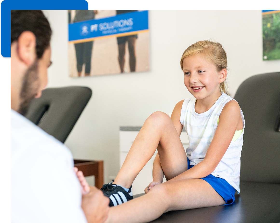 A pediatric physical therapist gently assessing a toddler's ankle range of motion on a therapy mat in a clinic, real photograph style