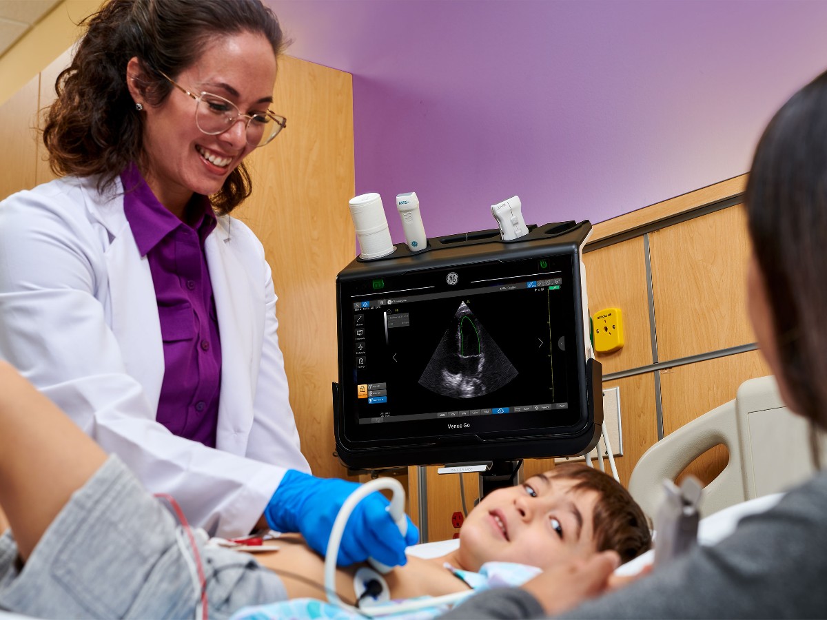 A pediatric sonographer performing an abdominal ultrasound on a toddler lying on a hospital bed in an emergency department, calm clinical setting, real-life photo