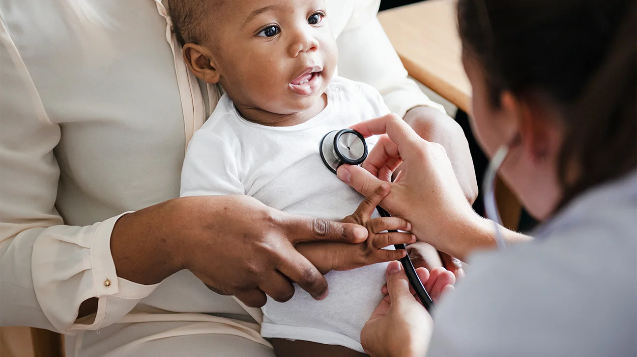 A pediatrician gently listening to a baby’s chest with a stethoscope in a bright exam room while a parent holds the baby, candid photorealistic healthcare photography
