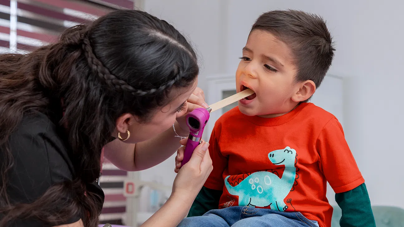 A pediatrician in a clinic gently examining a young child’s mouth with a small light while a parent stands close by, realistic medical photo