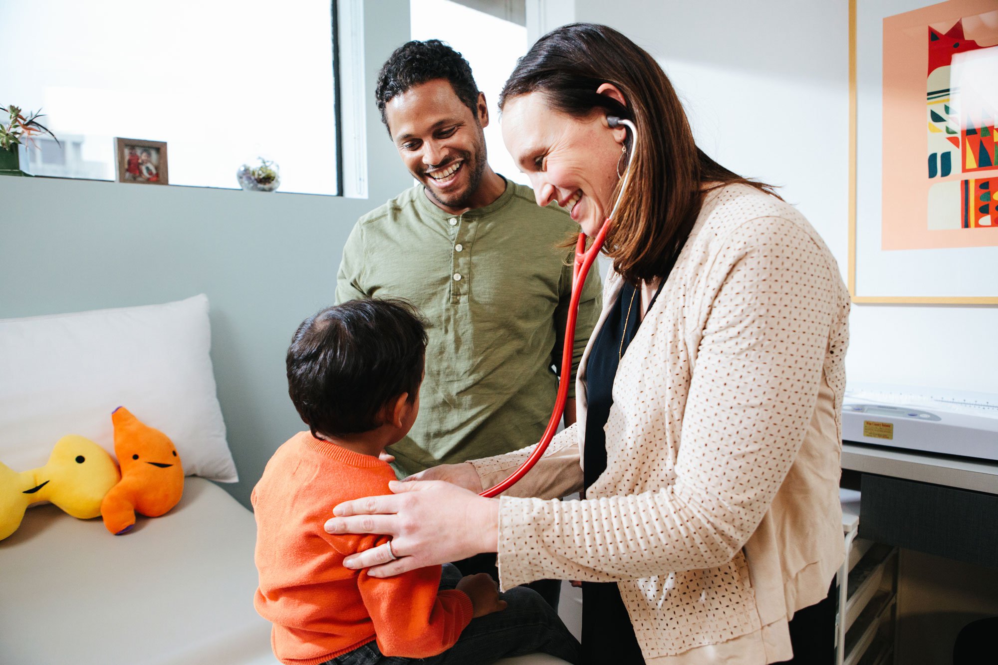 A pediatrician sitting in an exam room speaking with a parent while a toddler sits on the exam table, calm clinical setting, real photo style
