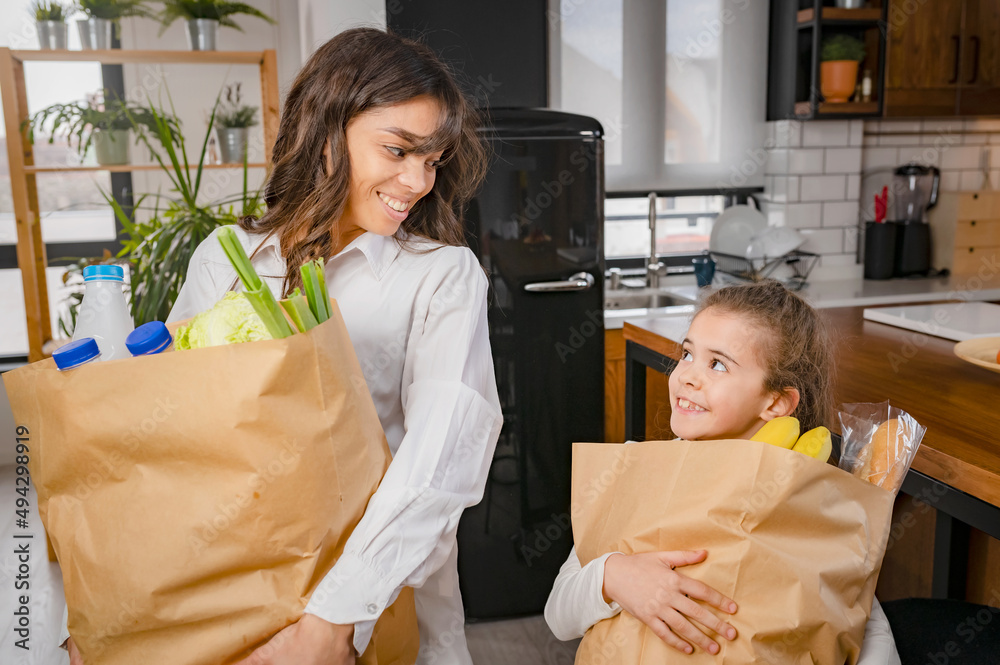 A preschool-aged child carrying a small grocery bag beside a parent in a kitchen, real-life supportive moment photo