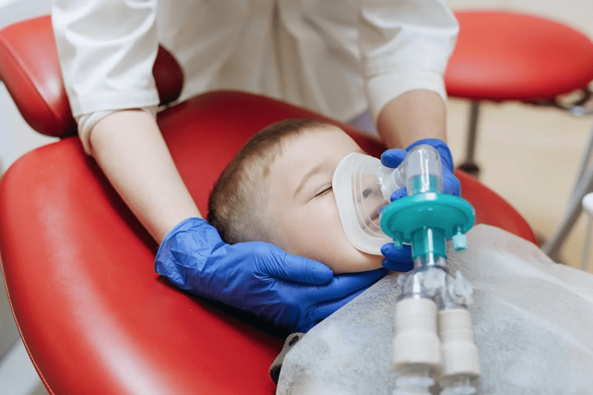 A preschool-aged child resting on a hospital recovery bed with a parent holding their hand, monitoring equipment in the background, outpatient surgery recovery photo