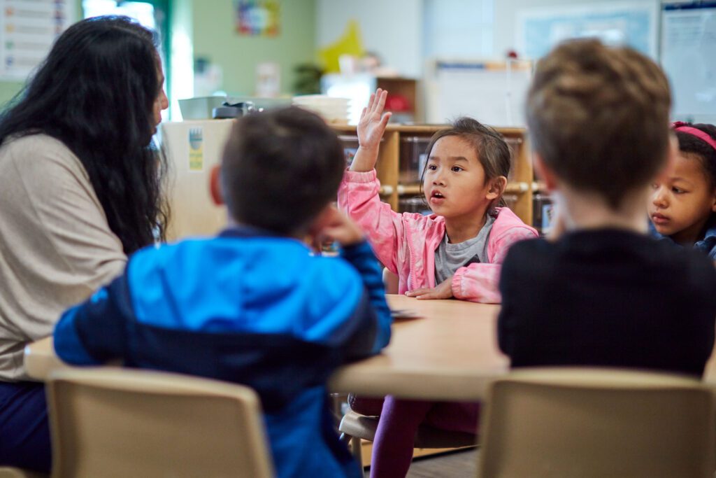 A preschool-aged child sitting at a classroom table leaning forward with a focused expression while a teacher speaks nearby, natural candid photo