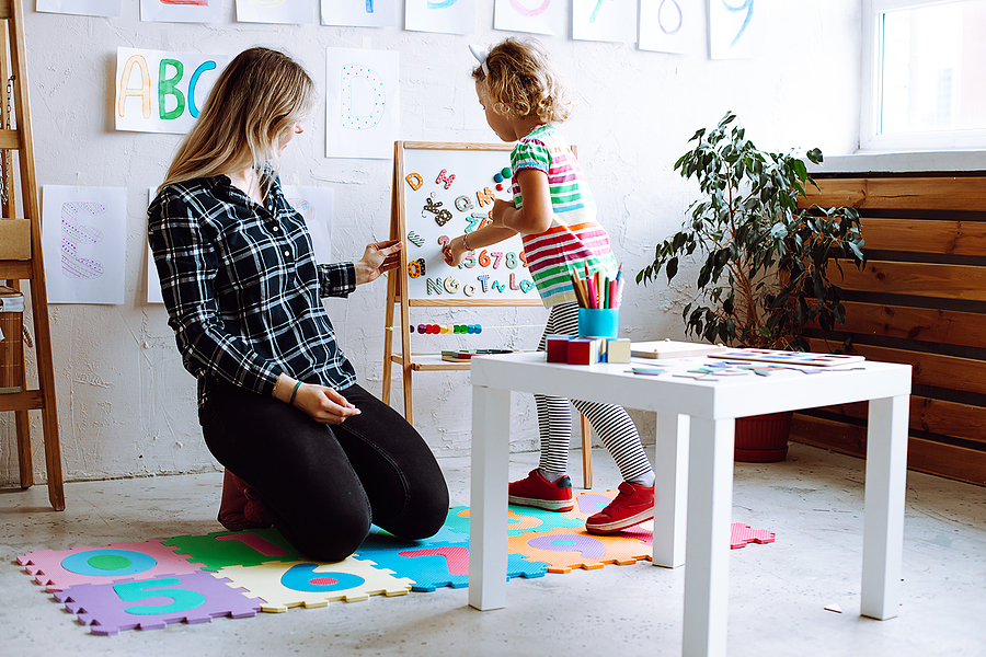 A preschool teacher kneeling beside a quiet child at a classroom table, offering a gentle smile and a supportive presence, candid photo