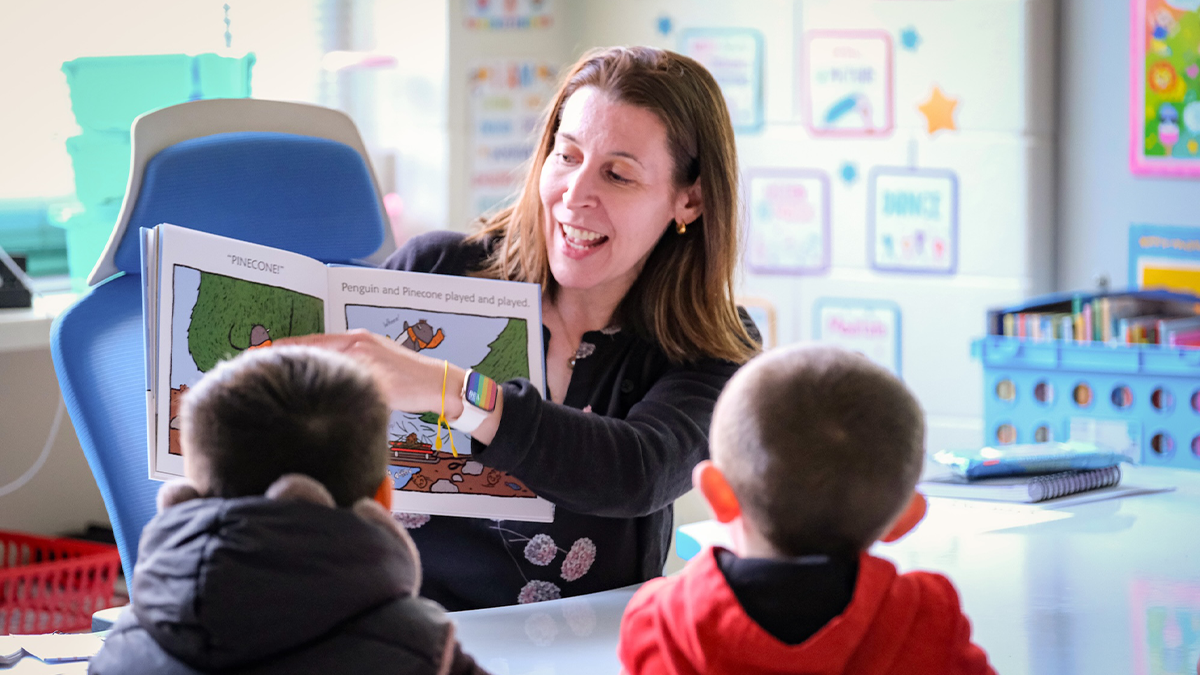 A preschool teacher sitting on a classroom rug reading a picture book to a small group of toddlers, candid classroom photo
