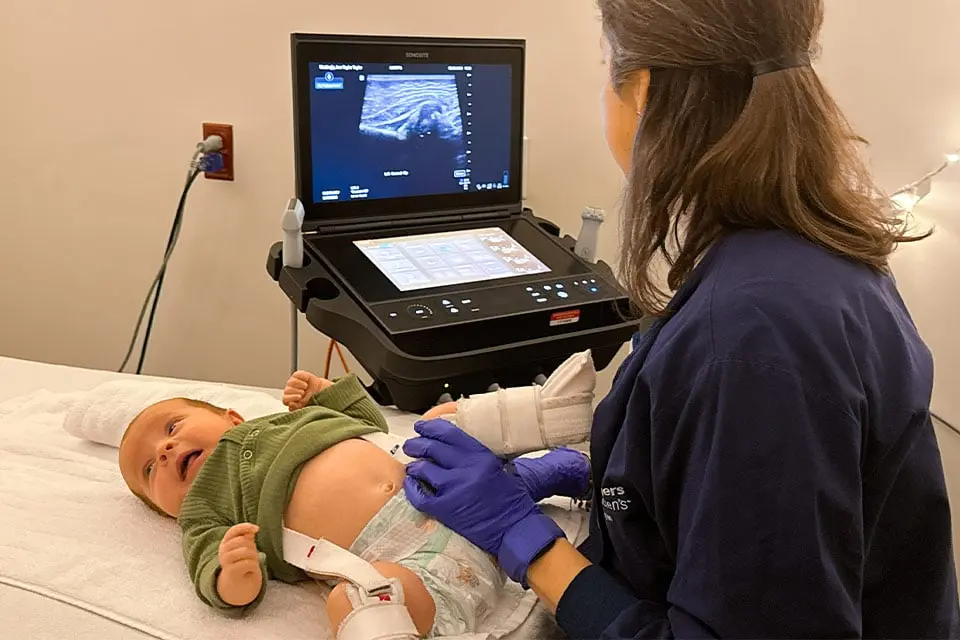 A radiology technician performing a hip ultrasound on an infant lying calmly on an exam table in a clinic room, realistic photo