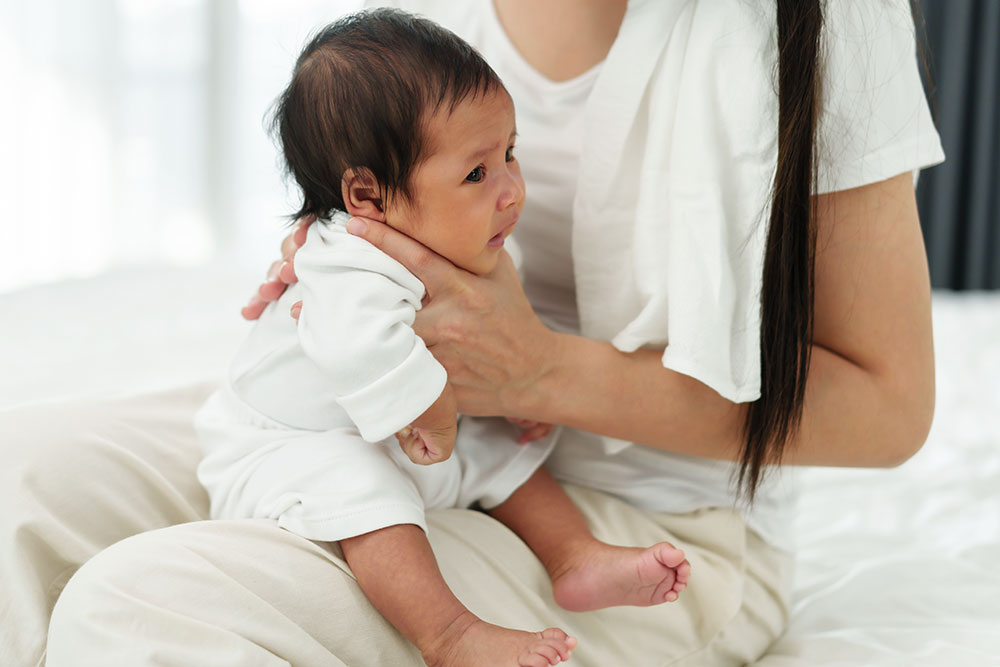 A real-life photo of a baby sitting on a parent’s lap in a supported seated burping position, with the parent’s hand supporting the baby’s chest and jaw while the other hand pats the baby’s back