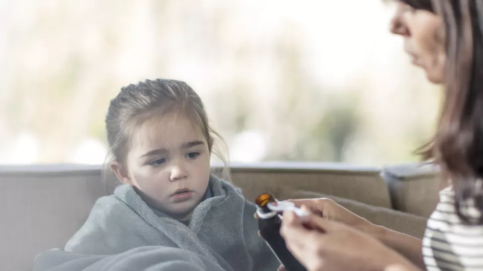A real-life photo of a parent gently giving liquid medicine with a syringe to a sleepy 15-month-old toddler in pajamas in a dim bedroom