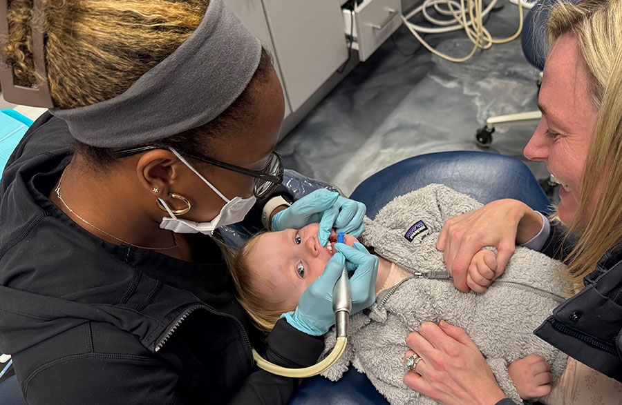 A real photo of a pediatric dentist wearing gloves gently examining a newborn’s mouth while the baby is cradled in a parent’s arms in a dental clinic