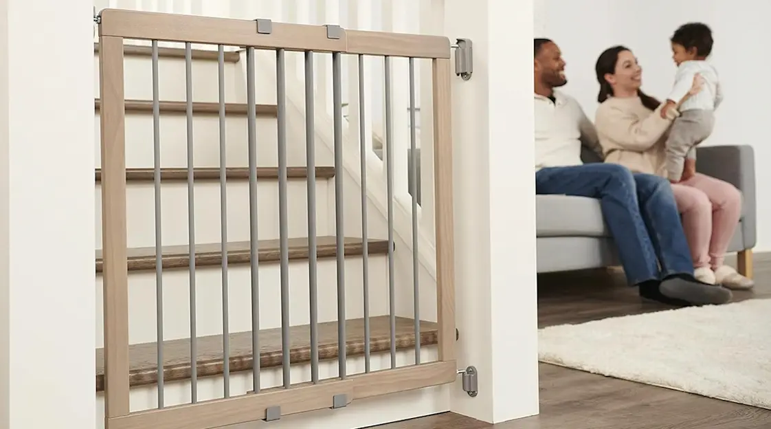 A real photograph of a hardware-mounted baby gate installed at the top of a staircase in a home hallway, neutral walls and natural light