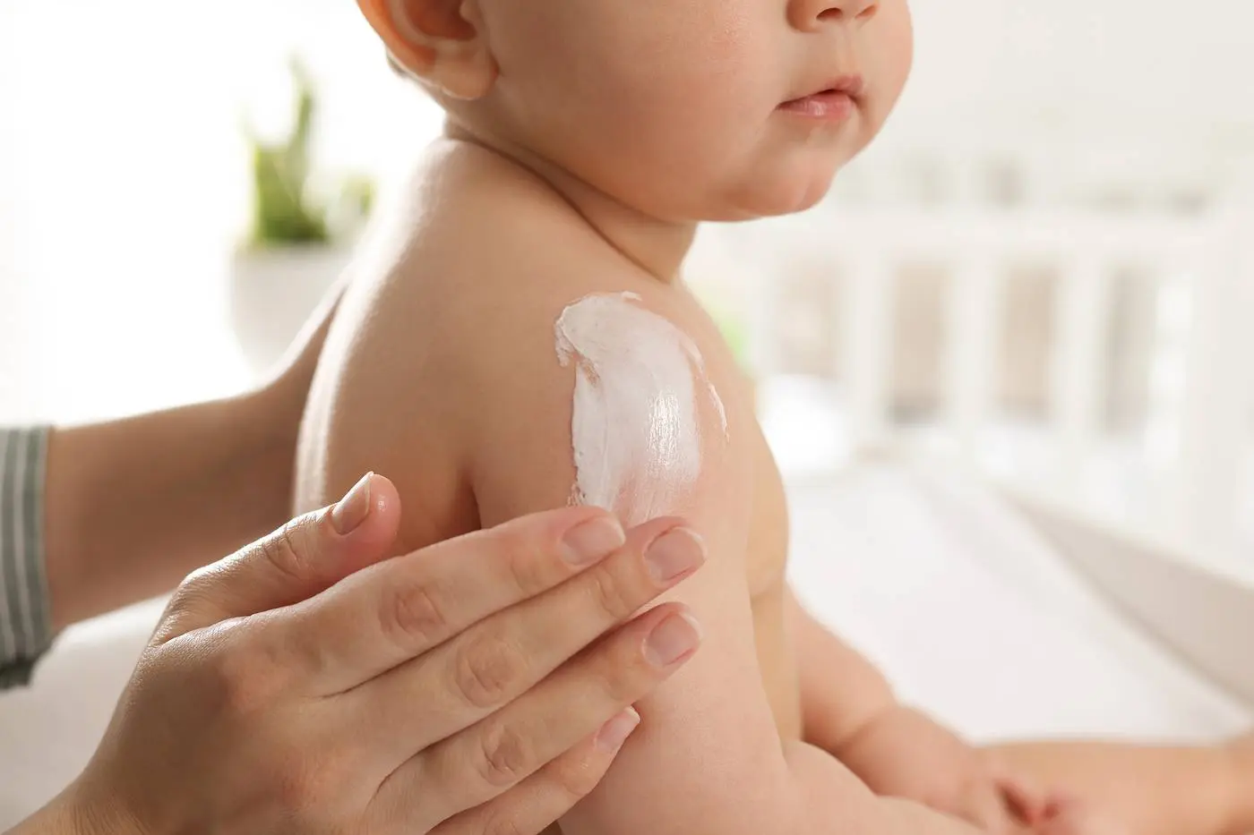 A real photograph of a parent’s hand applying a fragrance-free cream to a child’s upper arm in a bathroom with soft lighting
