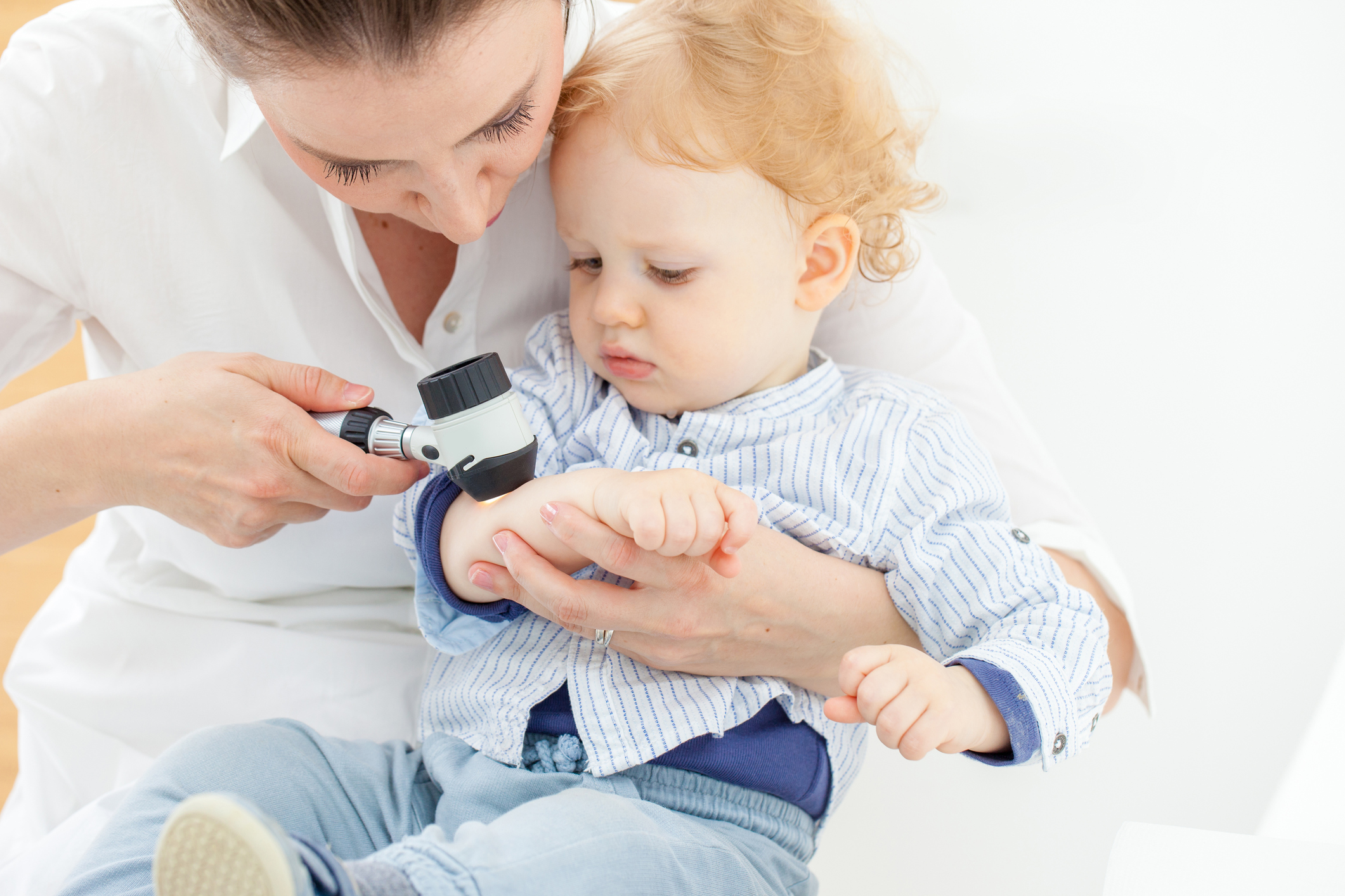 A real photograph of a pediatric dermatologist gently examining a baby’s skin on the shoulder in a clinic room while a parent holds the infant