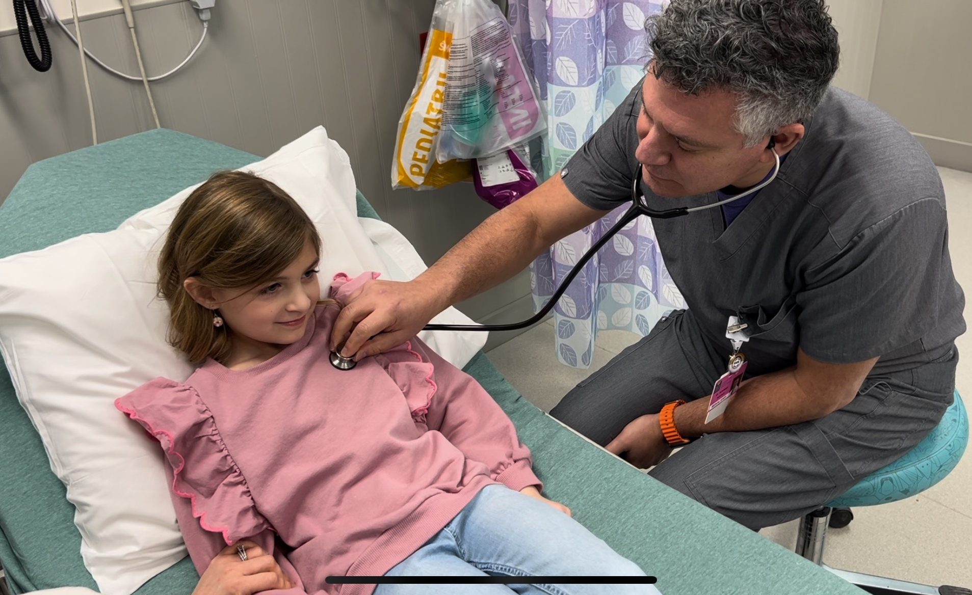 A real photograph of a pediatric triage nurse in an emergency department taking vital signs from a school-aged child seated on a hospital bed with a worried parent nearby