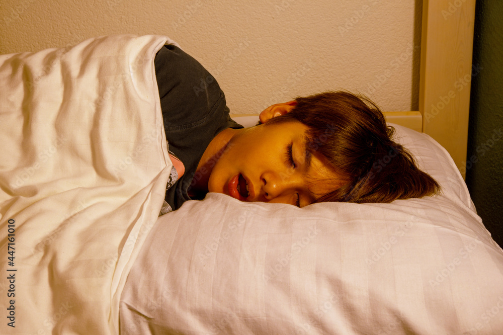 A school-age child asleep on a pillow in a softly lit bedroom, mouth slightly open as if snoring, candid photo