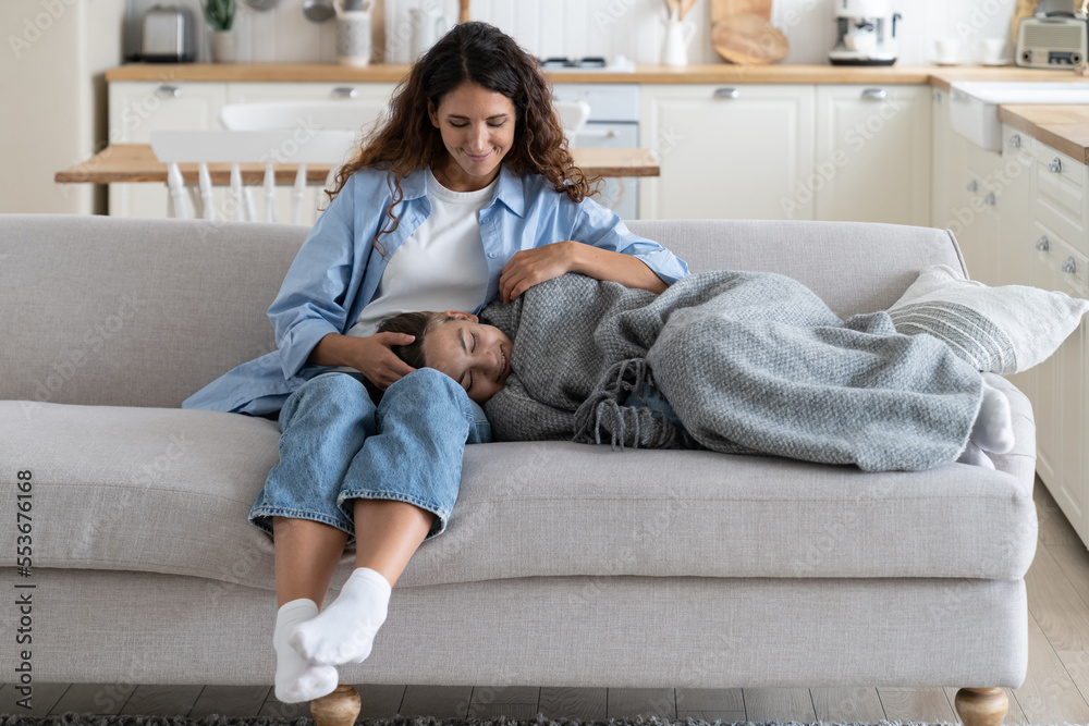 A school-age child lying on a living room couch under a blanket with a tired expression while a caregiver checks on them, natural indoor lighting, realistic photo