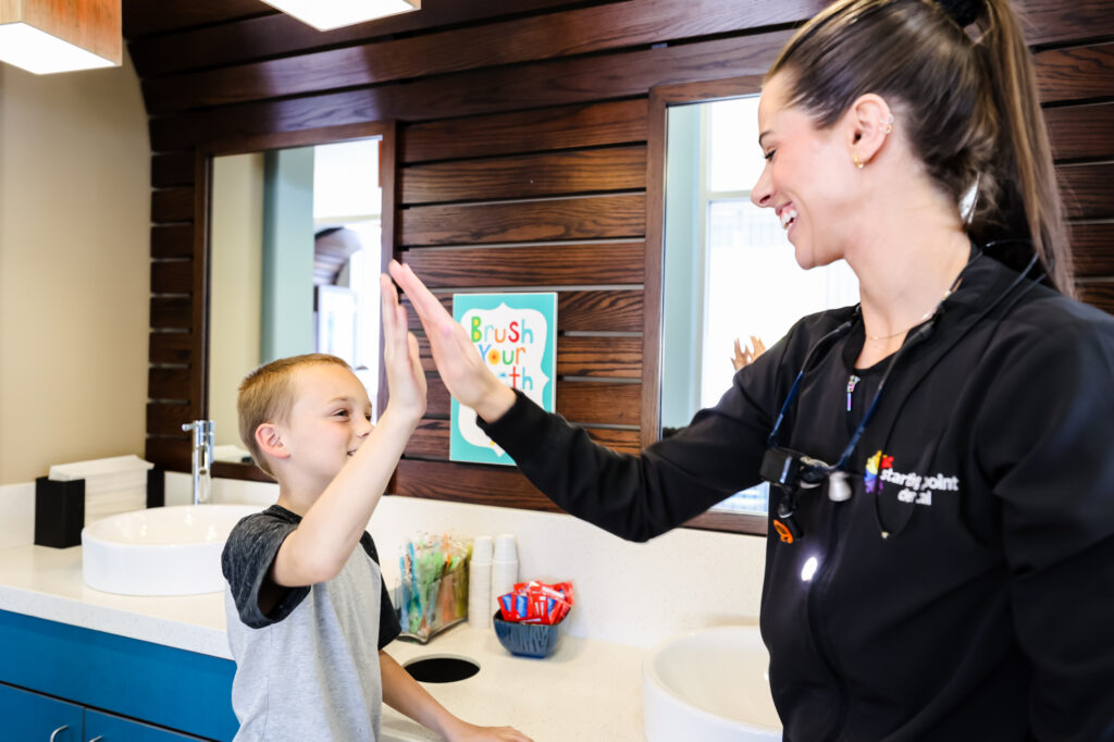 A school-aged child standing at a bathroom sink pointing to a back tooth while a parent looks on with concern, natural home lighting