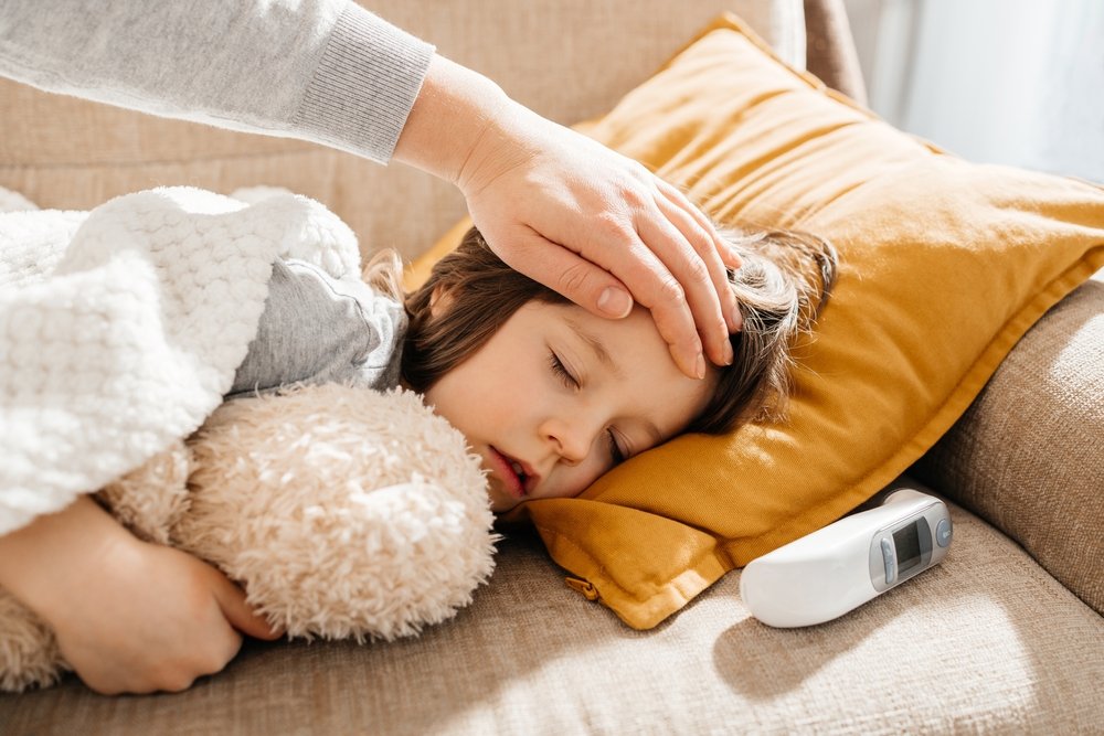 A sick toddler lying still on a couch with a blanket while a parent gently places a hand on the child's forehead, natural indoor light, realistic photo