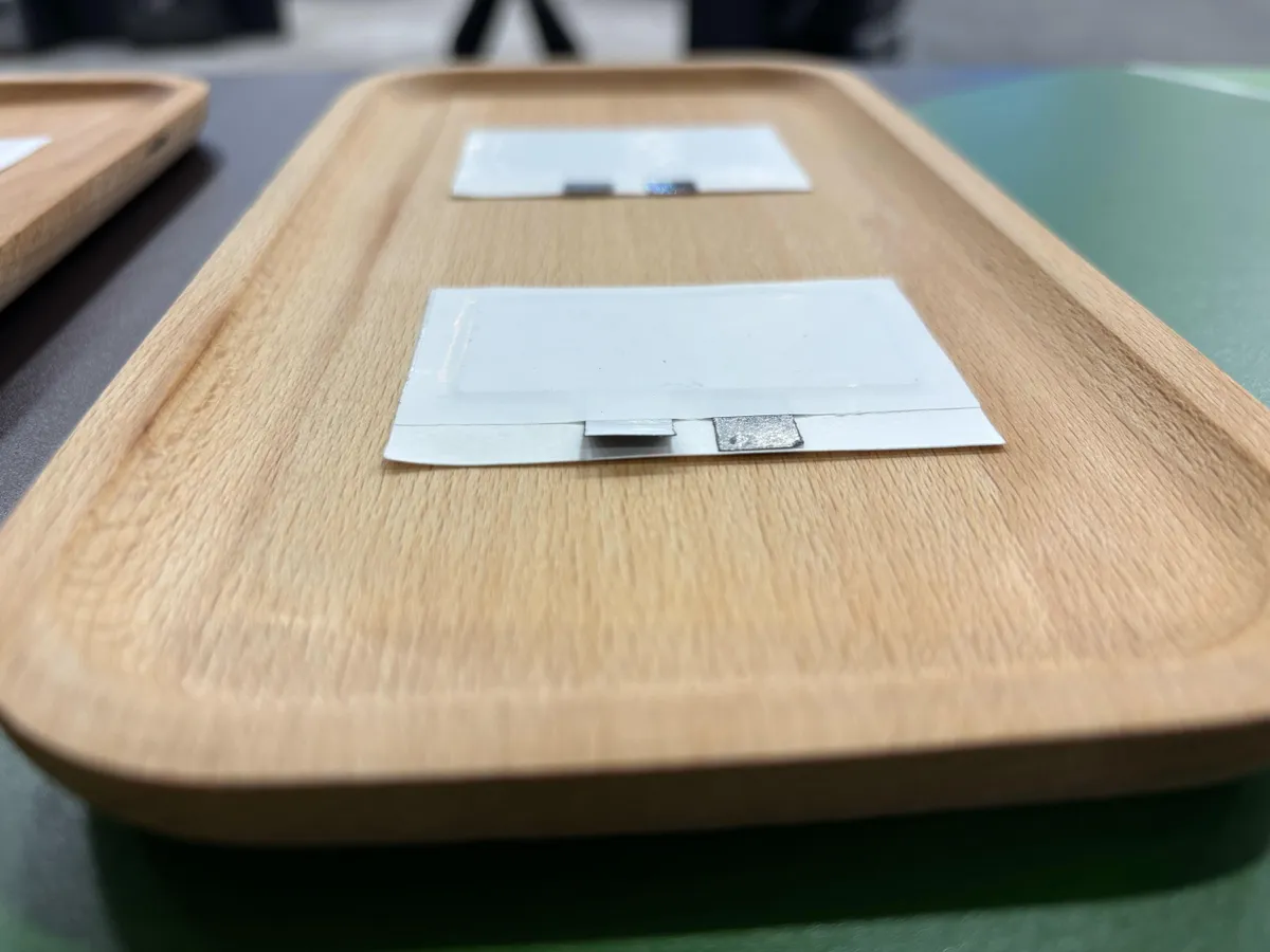 A single button battery resting on a wooden tabletop near a TV remote control, close-up photo with shallow depth of field