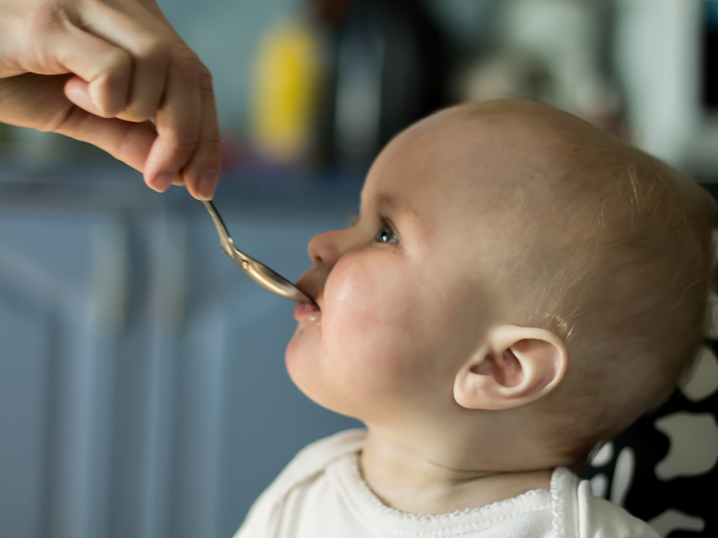 A sleepy newborn lying on a parent’s lap while a caregiver holds a small bottle of infant vitamin D drops above a clean teaspoon in soft natural window light, real photo