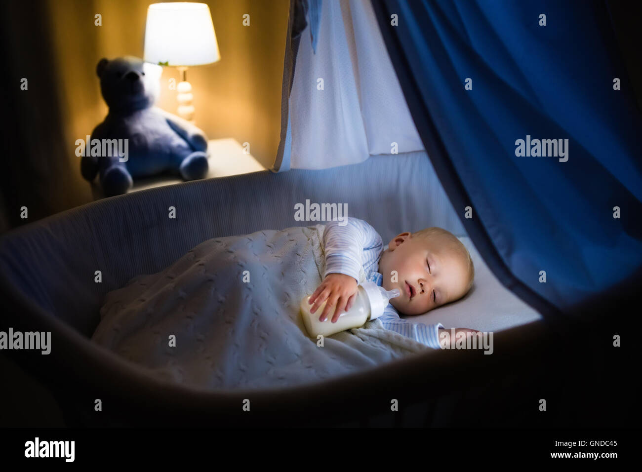 A sleepy toddler lying in a crib holding a baby bottle and drinking milk in a dimly lit bedroom at bedtime, natural photography style