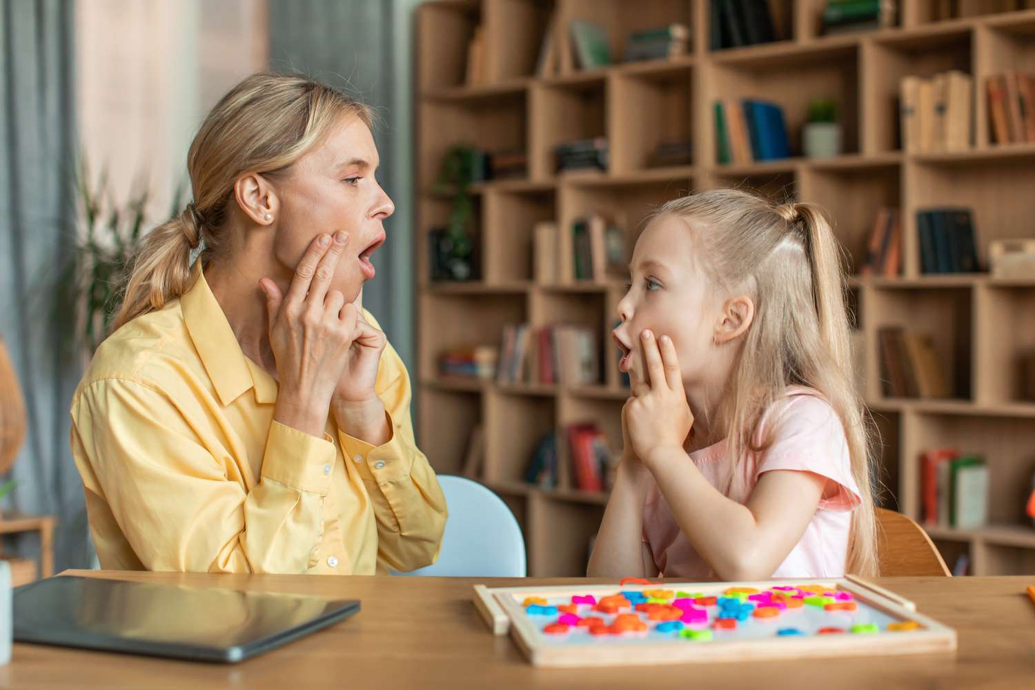 A speech-language pathologist kneeling beside a toddler and gently prompting them to imitate simple sounds using a toy and picture cards in a clinic setting
