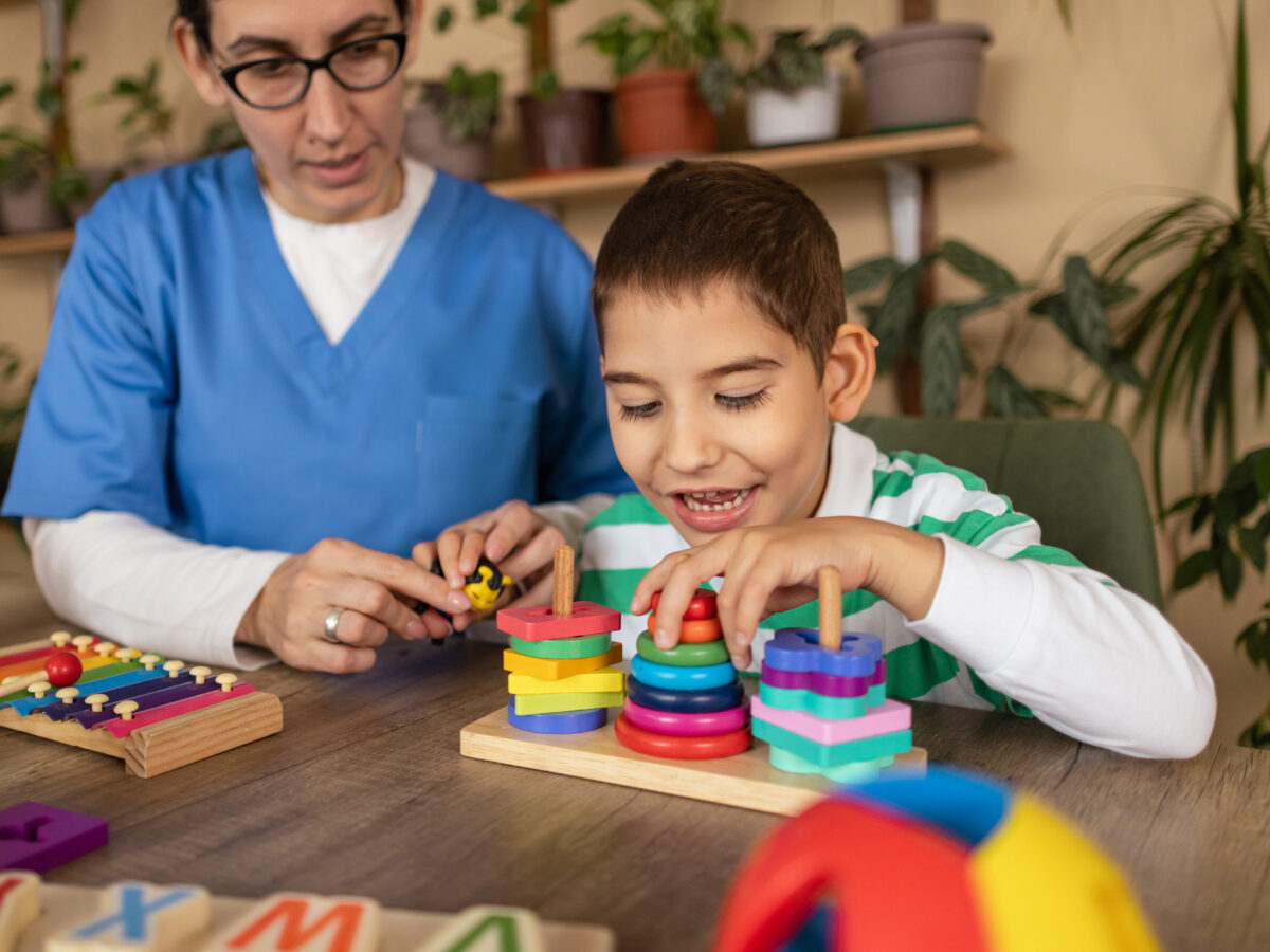 A speech therapist sitting on the floor in a pediatric clinic playroom building blocks with a toddler while the child vocalizes and gestures, candid photo