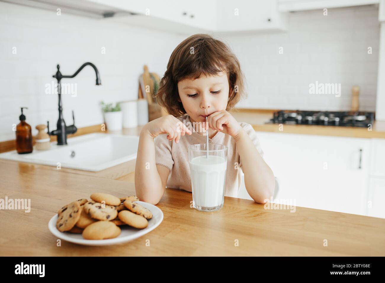 A tired-looking toddler sitting at a kitchen table holding a sippy cup of milk in warm morning light, real-life family photo style