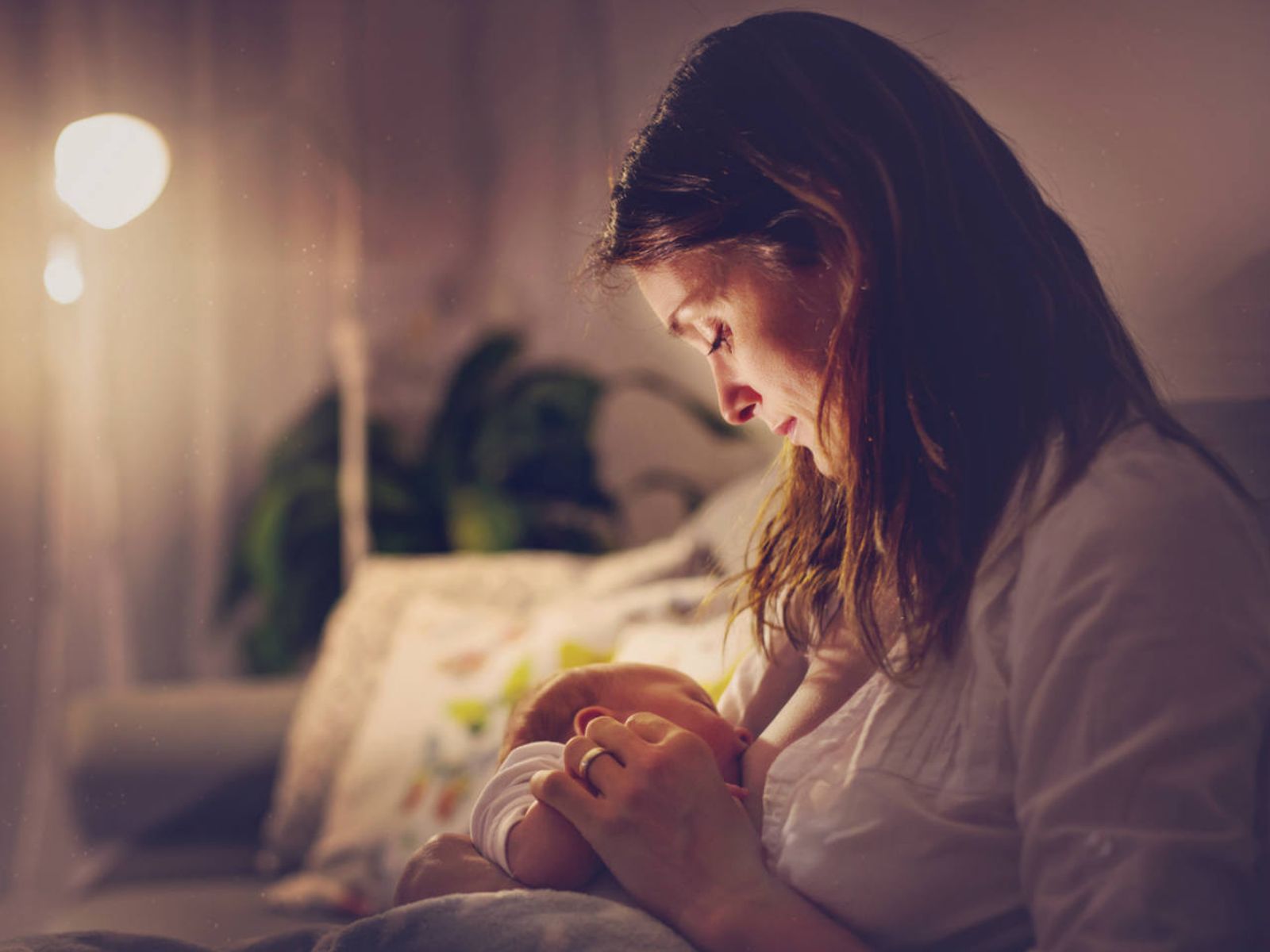 A tired mother sitting upright in bed breastfeeding a newborn in a dim bedroom at night with minimal bedding visible, realistic photo