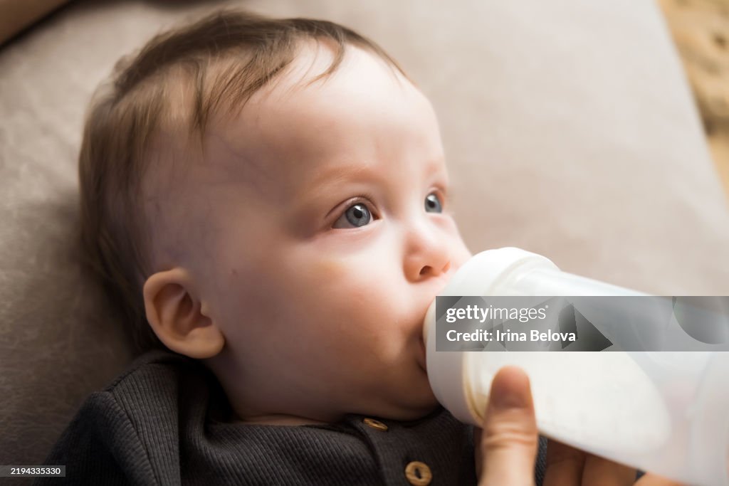 A tired parent holding a fussy infant who is slowly drinking from a baby bottle on a couch in soft natural window light