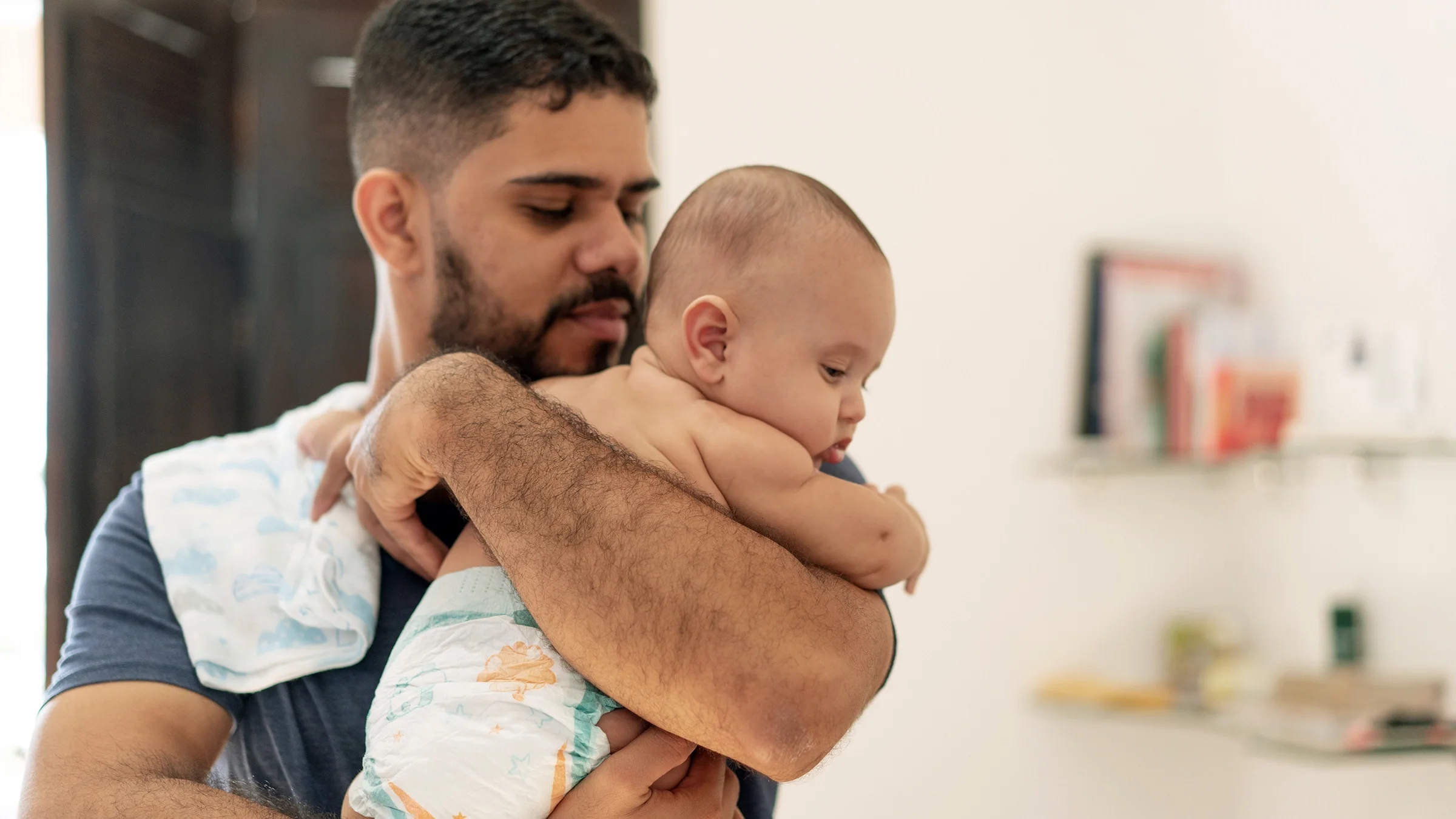 A tired parent holding a newborn baby upright against their chest in a softly lit living room, calming and supportive moment