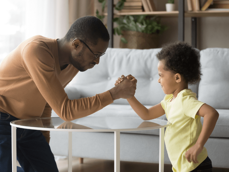 A tired parent kneeling in a living room, gently comforting an upset preschool-aged child who is crying near scattered toys, realistic family photograph