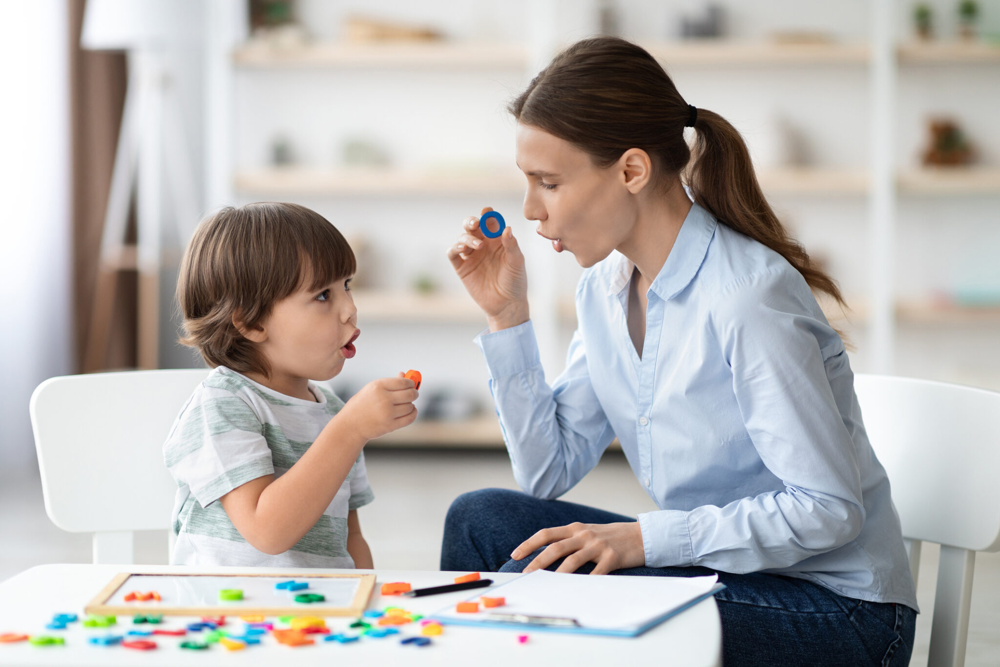 A tired parent sitting at a kitchen table holding a phone, with an 18-month-old toddler playing nearby, everyday home scene
