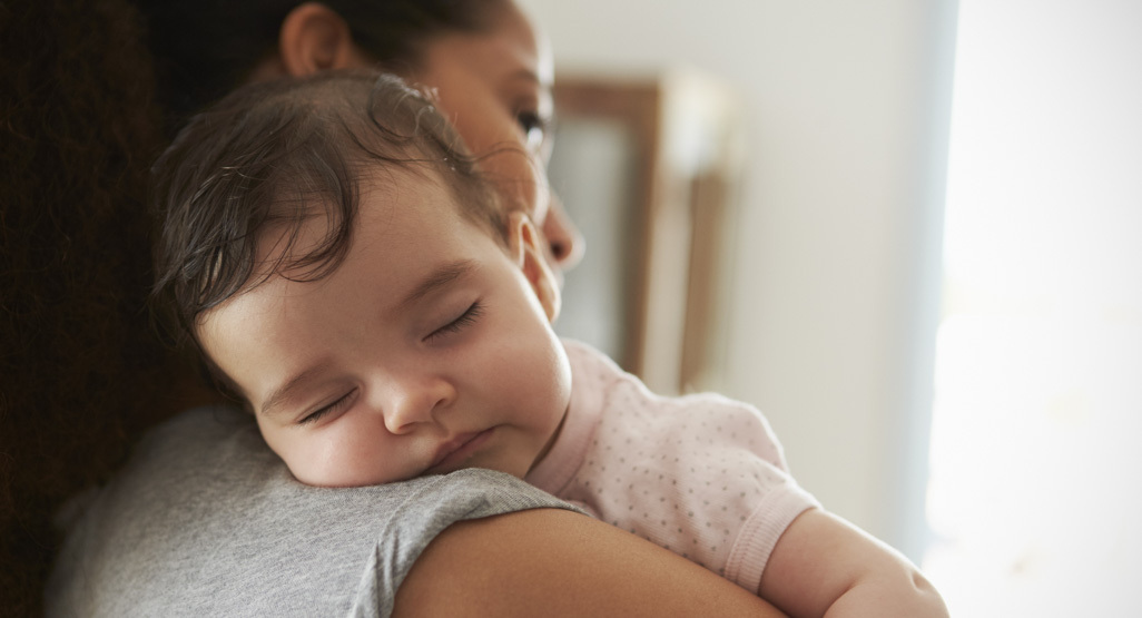 A tired parent sitting in a chair holding a sleeping seven-month-old baby for a daytime nap, soft window light, real photograph