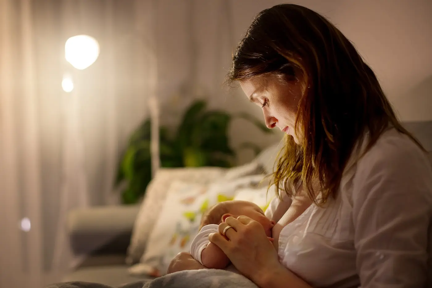 A tired parent sitting in a rocking chair at night holding a newborn against their chest, soft lamplight creating a calm, reassuring atmosphere