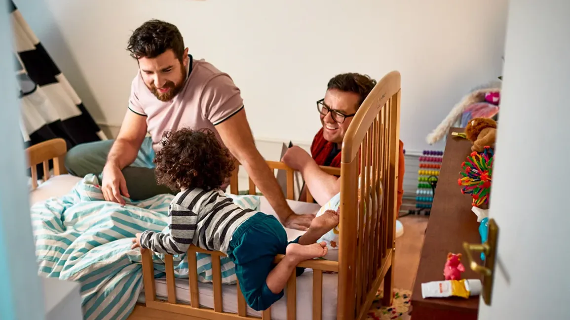 A tired parent sitting on a couch while a preschool-aged child bounces and climbs nearby during an evening routine, candid home photo