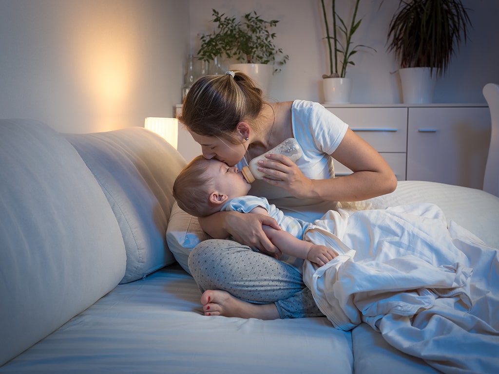 A tired parent sitting on the edge of a bed soothing a baby in a dark bedroom, realistic home photograph