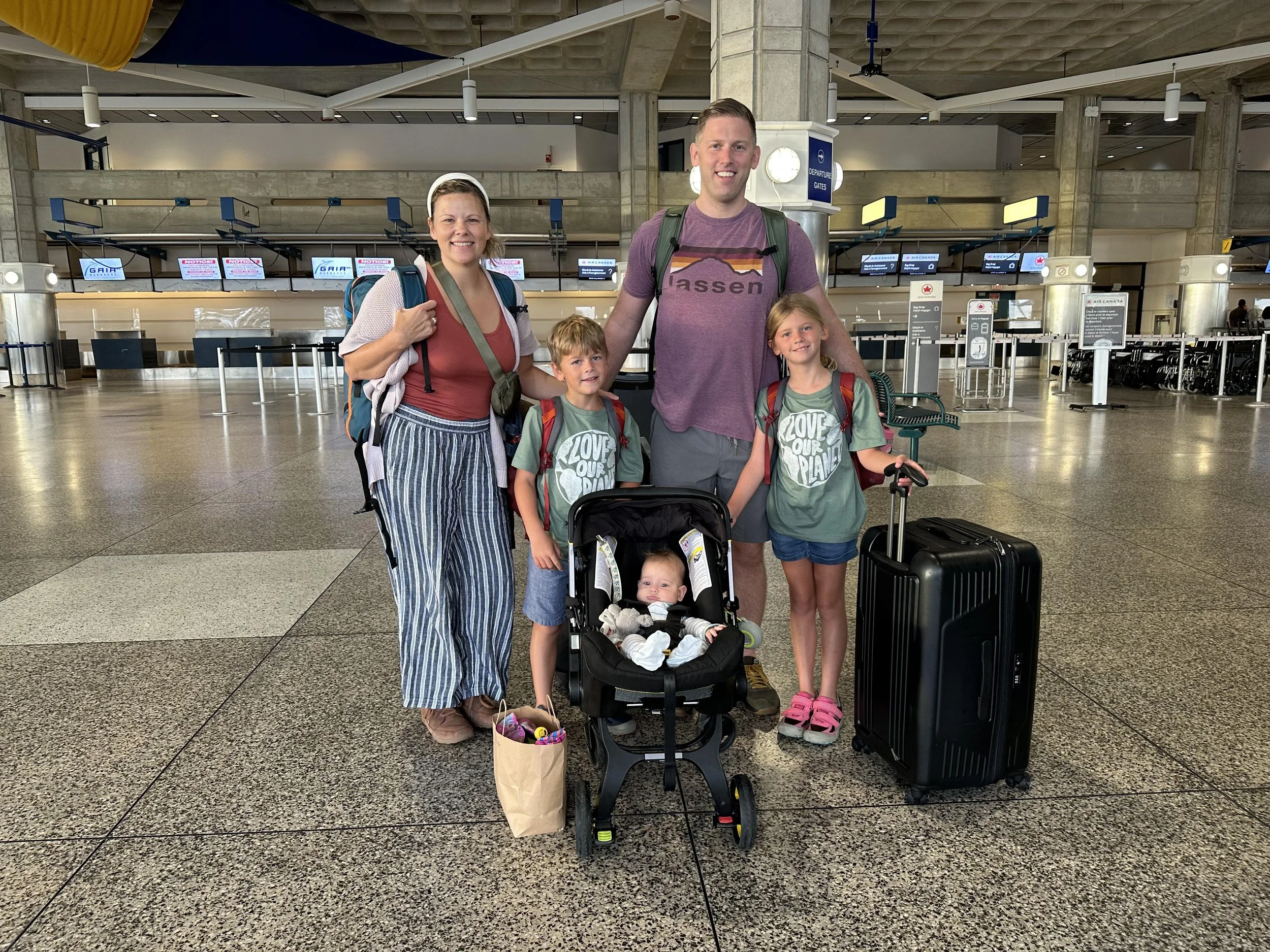 A tired parent wearing a baby in a soft carrier while standing in a bright airport terminal near large windows, real-life travel photo