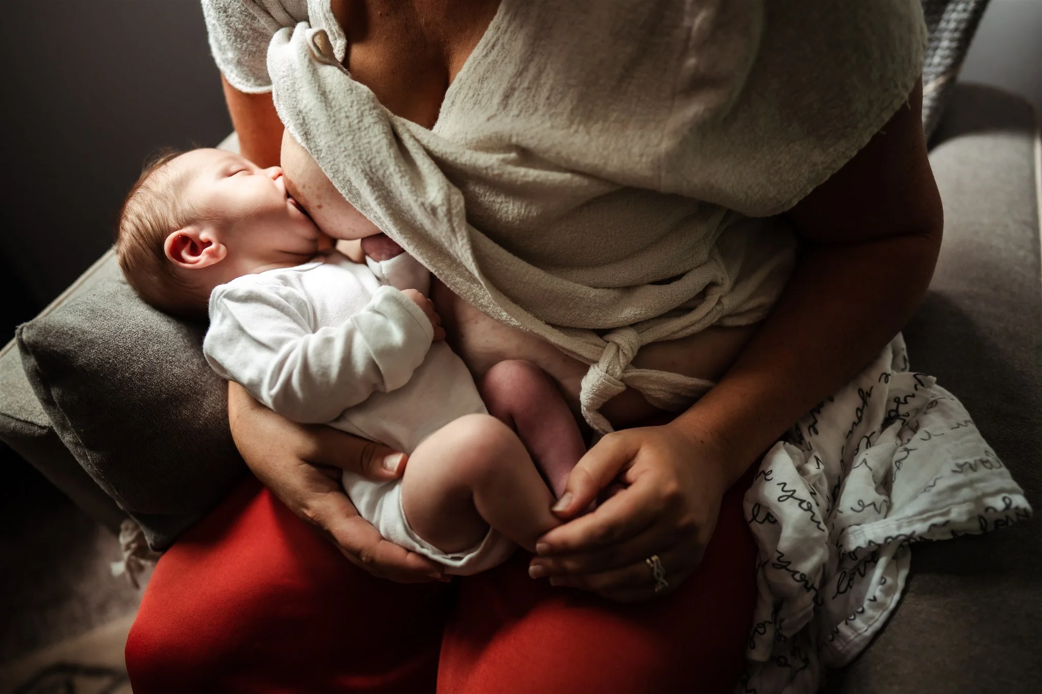 A tired postpartum mother sitting on a couch at night breastfeeding a newborn, warm lamp light in a calm living room, real-life photo style